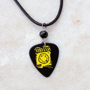 May include: Black guitar pick pendant necklace featuring the Nirvana logo in yellow on a black background. The pendant is attached to a black cord necklace with a black bead accent.