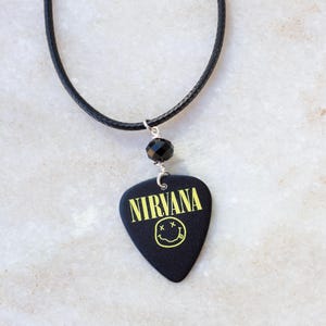May include: Black guitar pick necklace with a yellow Nirvana logo and smiley face. The necklace has a black cord and a black bead.