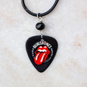 Puede incluir: Un collar con una púa de guitarra negra, una cuenta negra y un gráfico de lengua roja. La púa tiene el texto "The Rolling Stones Fifty Years".