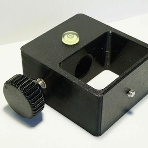 Pode incluir: Um suporte de montagem preto, retangular e impresso em 3D, com um botão de ajuste circular em um lado, um pequeno parafuso de metal na face frontal e um nível de bolha circular integrado na superfície superior para alinhamento preciso.