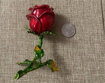 May include: A red rose with green leaves and gold accents, made of metal, is shown on a brown textured surface. The rose is about the size of a 20 pence coin.
