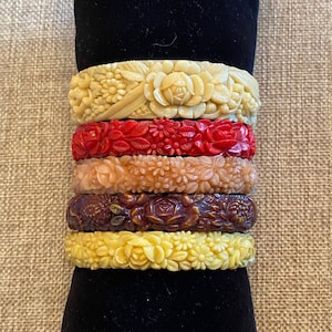 May include: Stack of six vintage plastic bangles with floral designs. The bangles are in various colors, including cream, red, peach, and brown.