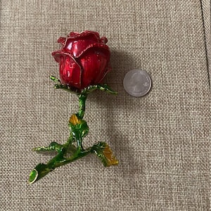 May include: A red rose with green leaves and gold accents, made of metal, is shown on a brown textured surface. The rose is about the size of a quarter.