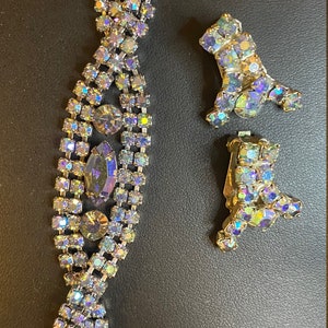 Op de afbeelding: Een set met iriserende strassjuwelen met een armband en een paar clip-on oorbellen. De armband is gemaakt van een ketting van kleine strasssteentjes en heeft een grotere, druppelvormige strasssteen in het midden. De oorbellen zijn ook gemaakt van strass en hebben de vorm van een druppel.