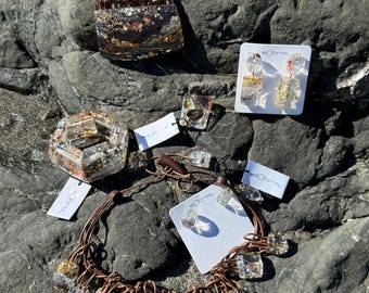 May include: A set of jewelry featuring a brown leather necklace with multiple clear resin pendants, a smaller clear resin pendant necklace, and a pair of clear resin earrings. The resin pieces have gold flecks and are displayed on a rocky surface.