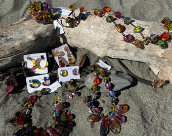 May include: A set of colorful glass necklaces and earrings. The necklaces are made of large, irregular shaped glass beads in shades of red, orange, yellow, green, blue, and purple. The earrings are made of smaller, round glass beads in the same colors. The jewelry is displayed on a piece of driftwood against a background of sand.