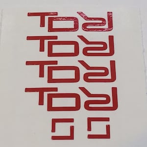 May include: Five red vinyl stickers with the word "TODJ" written in a stylized font. The stickers are arranged vertically, with the bottom sticker partially obscured by the edge of the image.