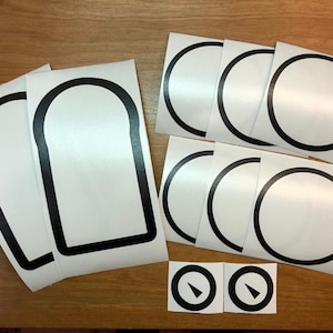 May include: A set of white vinyl stickers with black outlines. The set includes three rectangular stickers with rounded corners and five circular stickers. Two of the circular stickers have a black arrow pointing north.