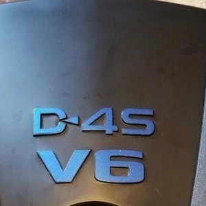 May include: Close-up of a black engine cover with a blue D-4S V6 emblem.