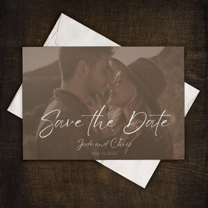 May include: A save the date card featuring a sepia-toned photograph of a couple embracing. The card includes the text "Save the Date" in elegant script, along with the names "Jade and Chris" and the date "May 10, 2024". The card is placed on a dark wooden surface.