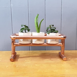 May include: Copper pipe plant stand with three small white pots and green plants. The stand has two shelves and a simple design.