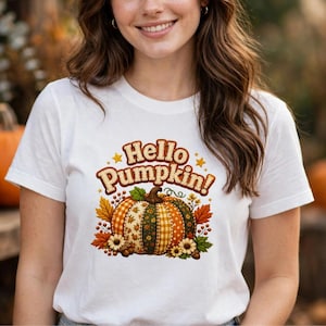 Puede incluir: Una camiseta blanca de manga corta con un estampado gráfico de una calabaza con varios patrones otoñales. El texto "Hello Pumpkin!" está impreso en marrón sobre la calabaza, rodeado de pequeñas estrellas amarillas, hojas naranjas y flores.