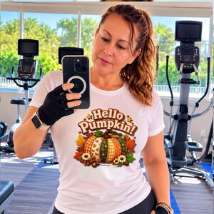 Puede incluir: Una camiseta blanca de manga corta con un estampado gráfico de una calabaza con múltiples patrones, hojas de otoño y flores. El texto "Hello Pumpkin!" está impreso en marrón sobre la calabaza. Tiene cuello redondo y corte ajustado.
