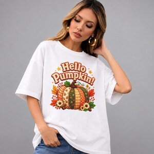 Puede incluir: Una camiseta blanca de manga corta con un estampado gráfico de una calabaza con patrón de patchwork en tonos naranja, verde y amarillo. El texto "Hello Pumpkin!" aparece impreso encima en una fuente marrón, redondeada y retro.