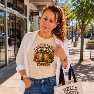 Puede incluir: Una camiseta de manga corta color crema con un gráfico de una calabaza estampada rodeada de hojas de otoño y girasoles. El texto dice Hello Pumpkin! TORVER! La camiseta se lleva debajo de una camisa blanca con pantalones vaqueros azules.