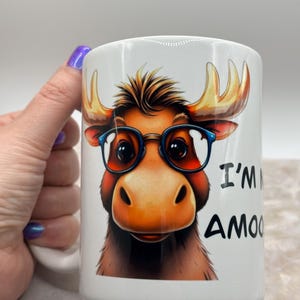 May include: White ceramic mug featuring a cartoon moose wearing blue glasses. The moose has brown fur and large antlers. Text on the mug reads "I'm Amoo".