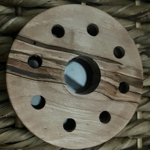 May include: A round wooden object with a central hole and eight smaller holes evenly spaced around the perimeter. The wood grain features a mix of light and dark brown tones. The object is likely a decorative element or part of a larger piece.