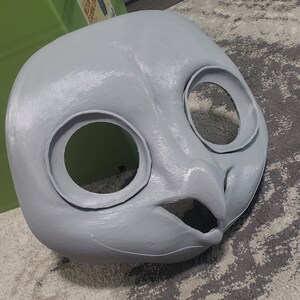 May include: A gray, unpainted, 3D printed animal mask with large eye holes and a beak. The mask is lying on a gray and white patterned rug.