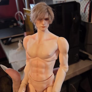 May include: A ball-jointed doll with light skin tone, defined muscular torso, and short, light brown hair. The doll features articulated joints at the shoulders, elbows, and wrists, with a neutral facial expression and detailed sculpted features.