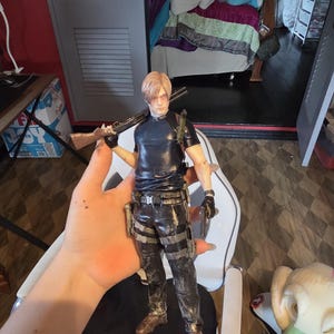 K&ouml;nnte beinhalten: Detaillierte Actionfigur eines m&auml;nnlichen Charakters, der eine Schrotflinte und eine Handfeuerwaffe h&auml;lt. Die Figur tr&auml;gt ein schwarzes Hemd, taktische Hosen und braune Stiefel. Die Figur ist etwa 30 cm gro&szlig;.