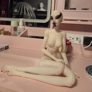 K&ouml;nnte beinhalten: Eine Puppe mit blasser Haut, schwarzem Augen-Make-up und einem schwarzen Lippen-Design sitzt auf einer rosa Oberfl&auml;che. Die Puppe hat spitze Ohren und schwarzen Nagellack. Die Puppe ist so positioniert, dass ein Bein &uuml;ber dem anderen gekreuzt ist.