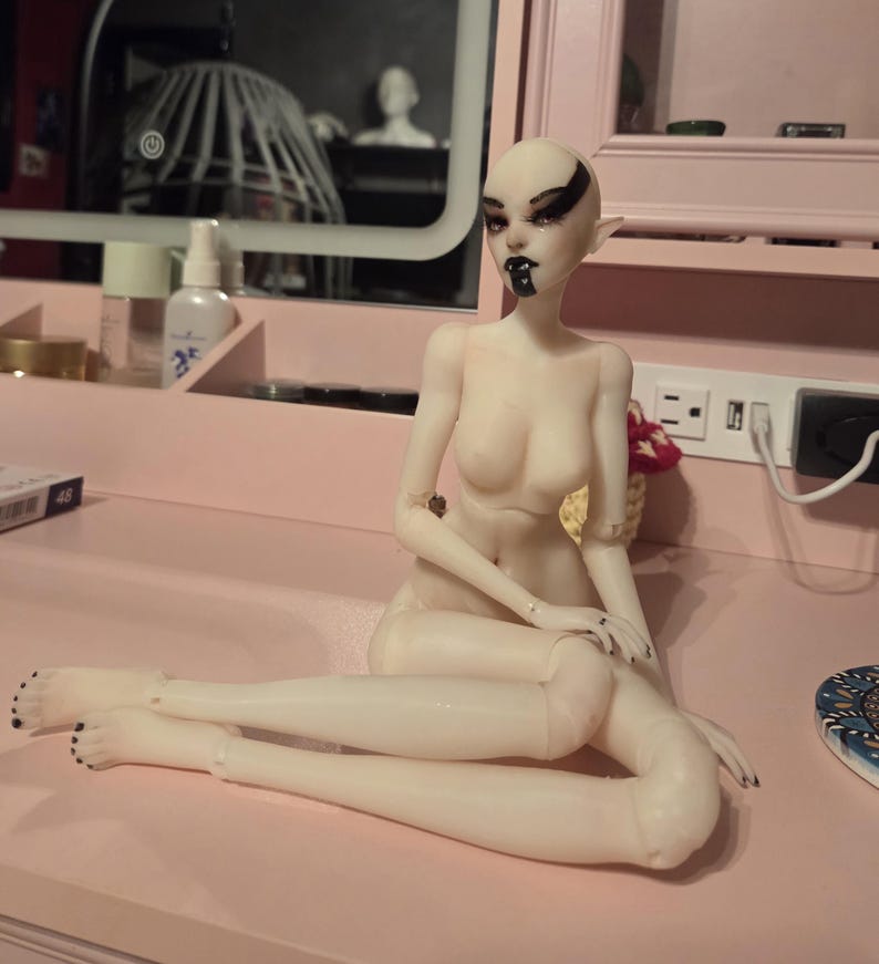 K&ouml;nnte beinhalten: Eine Puppe mit blasser Haut, schwarzem Augen-Make-up und einem schwarzen Lippen-Design sitzt auf einer rosa Oberfl&auml;che. Die Puppe hat spitze Ohren und schwarzen Nagellack. Die Puppe ist so positioniert, dass ein Bein &uuml;ber dem anderen gekreuzt ist.