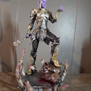 K&ouml;nnte beinhalten: Detaillierte Figur einer Fantasy-Figur in aufwendiger Gold- und Silberr&uuml;stung, mit violetter Haut und wei&szlig;em Haar. Die Figur steht auf einem Sockel mit einem erschlagenen Tierkopf, der von Pfeilen durchbohrt ist. Die Figur h&auml;lt eine Keule.
