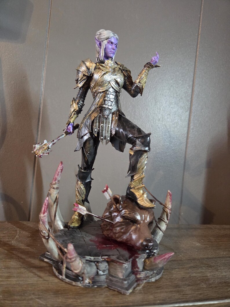 K&ouml;nnte beinhalten: Detaillierte Figur einer Fantasy-Figur in aufwendiger Gold- und Silberr&uuml;stung, mit violetter Haut und wei&szlig;em Haar. Die Figur steht auf einem Sockel mit einem erschlagenen Tierkopf, der von Pfeilen durchbohrt ist. Die Figur h&auml;lt eine Keule.