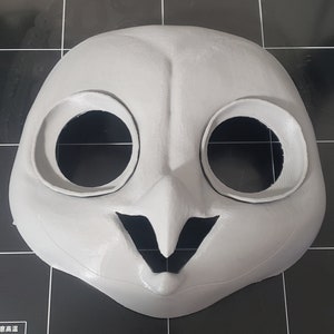 May include: A white 3D printed mask of an owl with large black eye holes and a black beak. The mask is on a black surface.