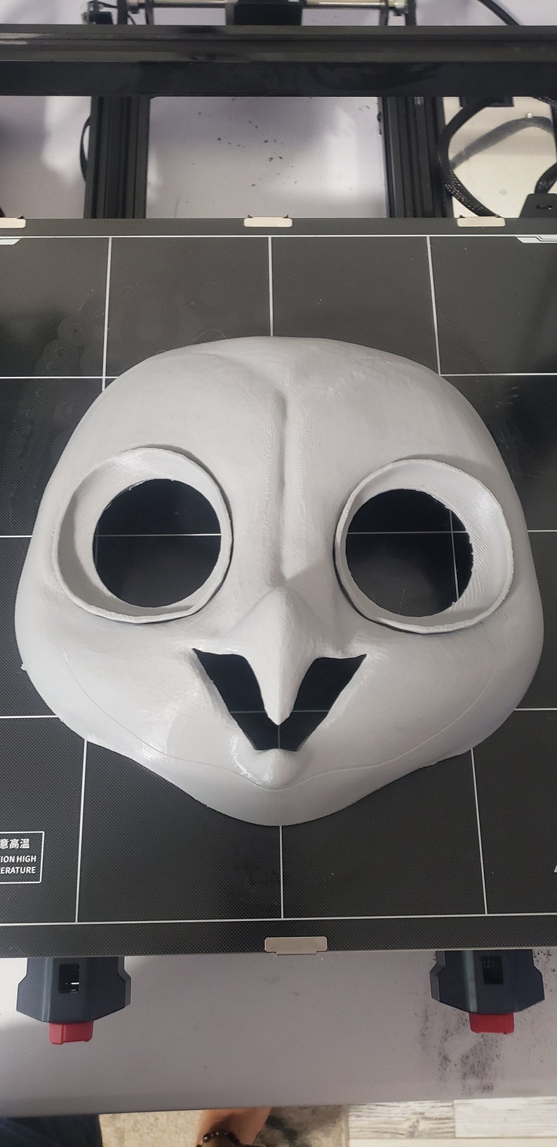 May include: A white 3D printed mask of an owl with large black eye holes and a black beak. The mask is on a black surface.