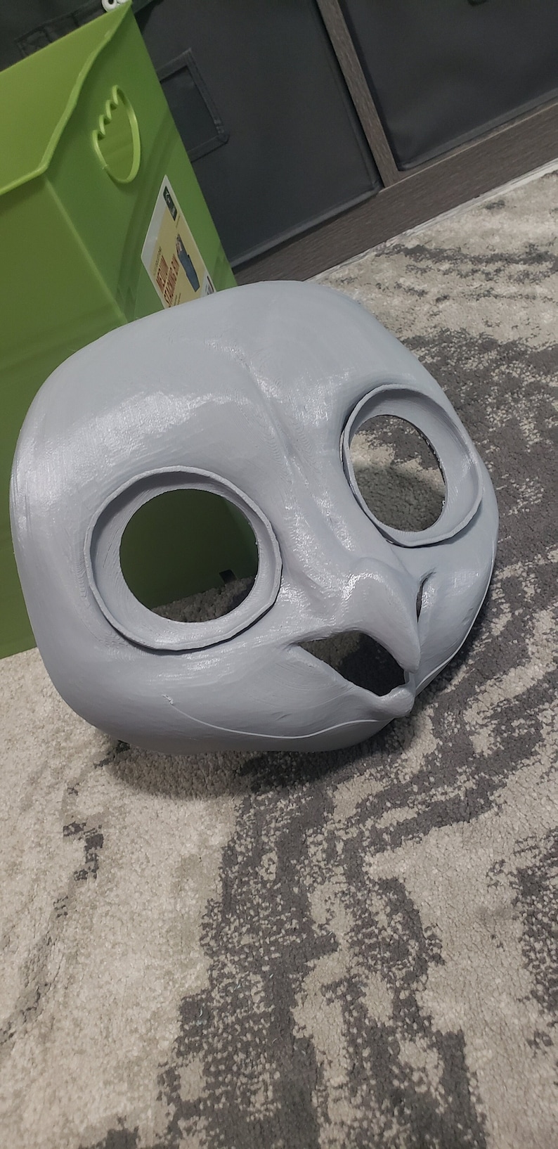 May include: A gray, unpainted, 3D printed animal mask with large eye holes and a beak. The mask is lying on a gray and white patterned rug.