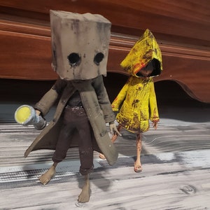 K&ouml;nnte beinhalten: Zwei detaillierte Figuren aus dem Videospiel Little Nightmares. Eine Figur tr&auml;gt eine braune Papiert&uuml;te &uuml;ber dem Kopf und einen langen Mantel und h&auml;lt eine Taschenlampe. Die andere Figur tr&auml;gt einen gelben Regenmantel.