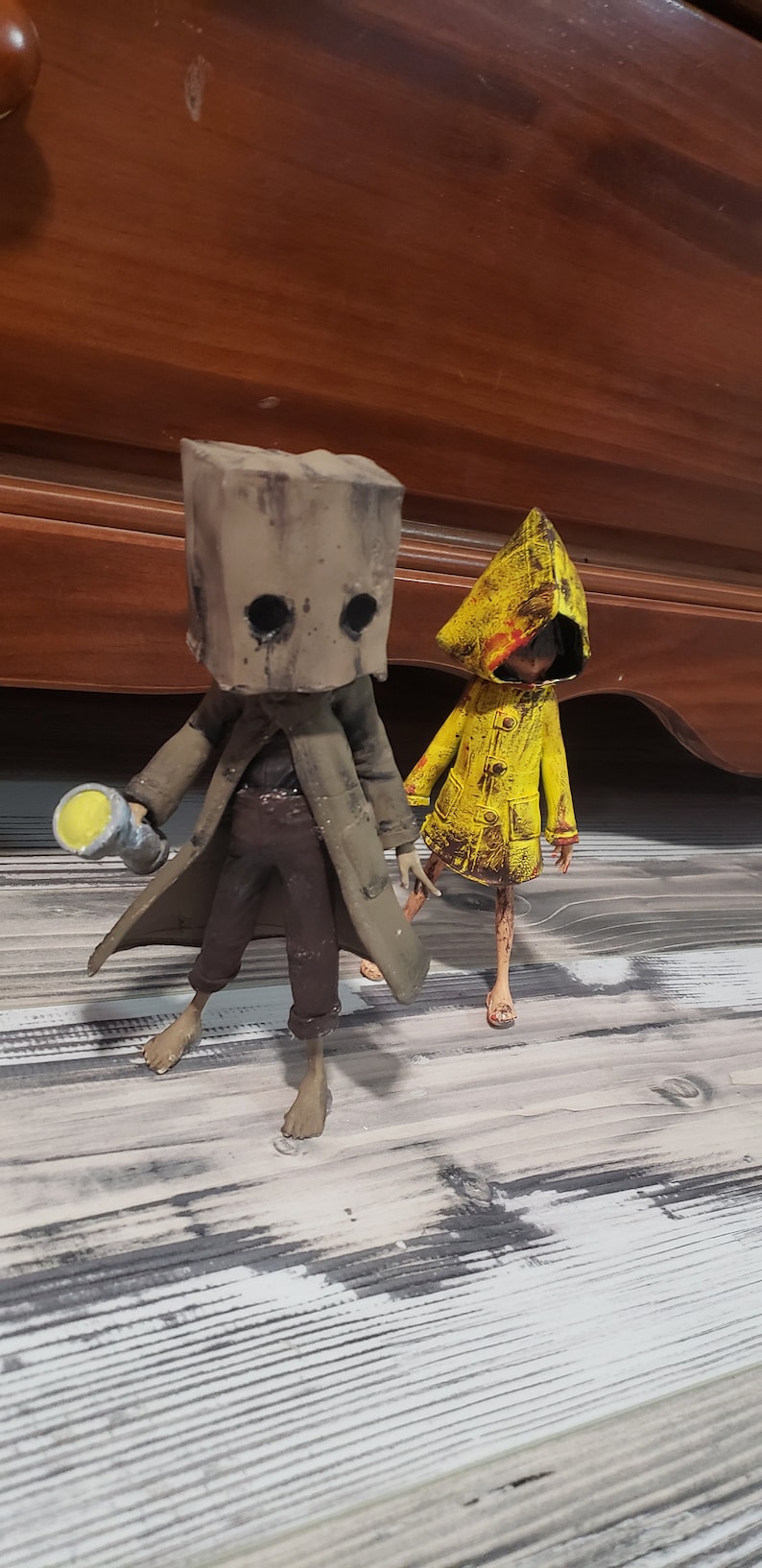 K&ouml;nnte beinhalten: Zwei detaillierte Figuren aus dem Videospiel Little Nightmares. Eine Figur tr&auml;gt eine braune Papiert&uuml;te &uuml;ber dem Kopf und einen langen Mantel und h&auml;lt eine Taschenlampe. Die andere Figur tr&auml;gt einen gelben Regenmantel.