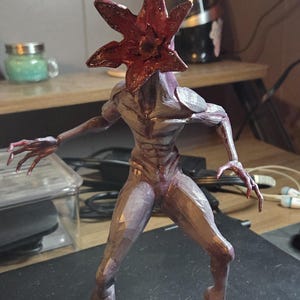 K&ouml;nnte beinhalten: Detaillierte Figur eines Demogorgon, einer Kreatur aus der Serie "Stranger Things". Die Figur hat einen rotbraunen, bl&uuml;tenblattartigen Kopf und einen humanoiden K&ouml;rper mit einem violett-grauen Farbschema. Die Figur ist mit ausgestreckten Armen posiert.