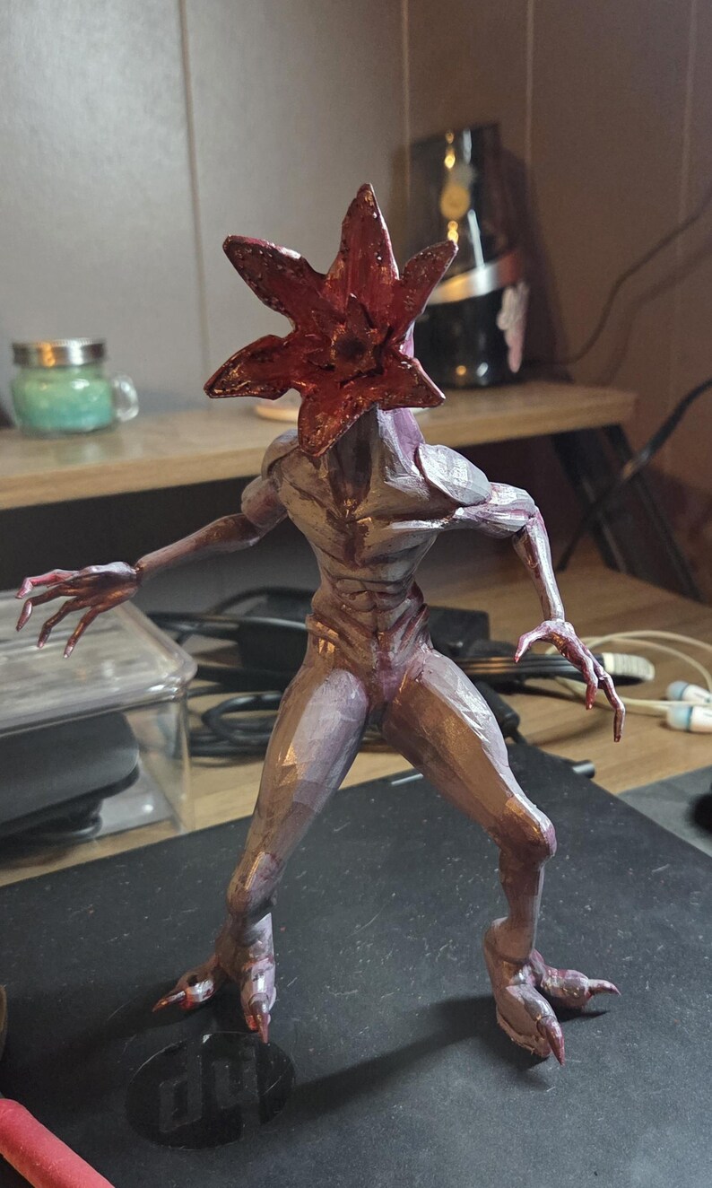 K&ouml;nnte beinhalten: Detaillierte Figur eines Demogorgon, einer Kreatur aus der Serie "Stranger Things". Die Figur hat einen rotbraunen, bl&uuml;tenblattartigen Kopf und einen humanoiden K&ouml;rper mit einem violett-grauen Farbschema. Die Figur ist mit ausgestreckten Armen posiert.