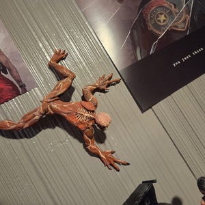 K&ouml;nnte beinhalten: Detaillierte, rotbraune Actionfigur einer Kreatur mit freiliegenden Muskeln und einem skelettartigen Brustkorb, dynamisch posiert. Die Figur befindet sich auf einer grauen Oberfl&auml;che, in der N&auml;he von Bildern einer Frau in einem roten Kleid und dem Text "you just think you have."