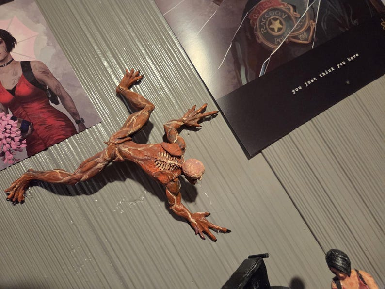 K&ouml;nnte beinhalten: Detaillierte, rotbraune Actionfigur einer Kreatur mit freiliegenden Muskeln und einem skelettartigen Brustkorb, dynamisch posiert. Die Figur befindet sich auf einer grauen Oberfl&auml;che, in der N&auml;he von Bildern einer Frau in einem roten Kleid und dem Text "you just think you have."