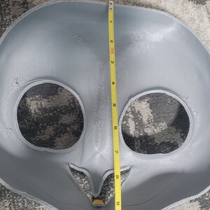 May include: A gray, unpainted, plastic mask with two eye holes and a mouth hole. The mask is being measured with a yellow tape measure, which shows a measurement of 10 inches.