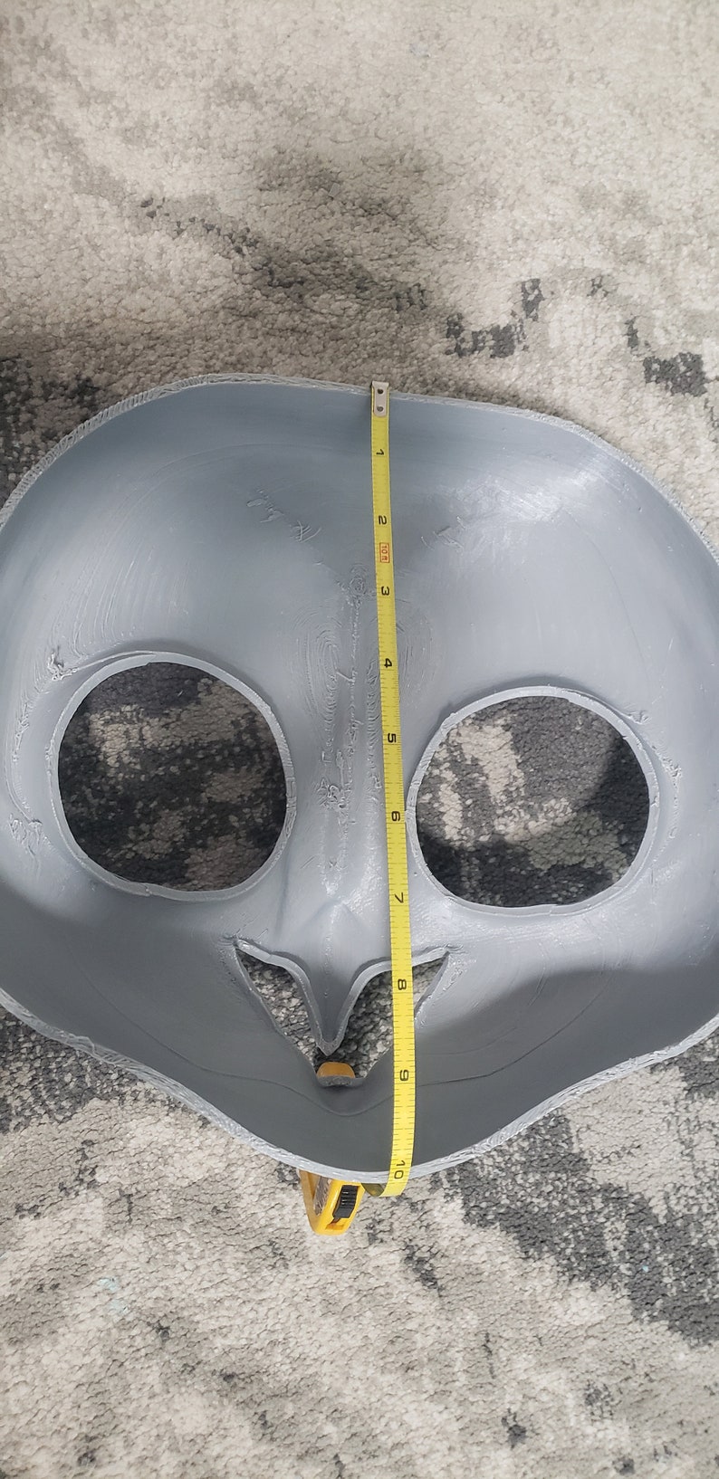 May include: A gray, unpainted, plastic mask with two eye holes and a mouth hole. The mask is being measured with a yellow tape measure, which shows a measurement of 10 inches.