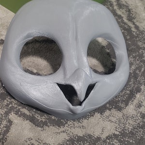 May include: A gray, unpainted, animal mask with large eye holes and a small mouth opening. The mask is made of a smooth, hard material.