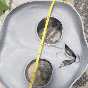 May include: A gray plaster mold of a face with two eye holes and a mouth hole. A yellow measuring tape is wrapped around the mold, measuring 10 inches.