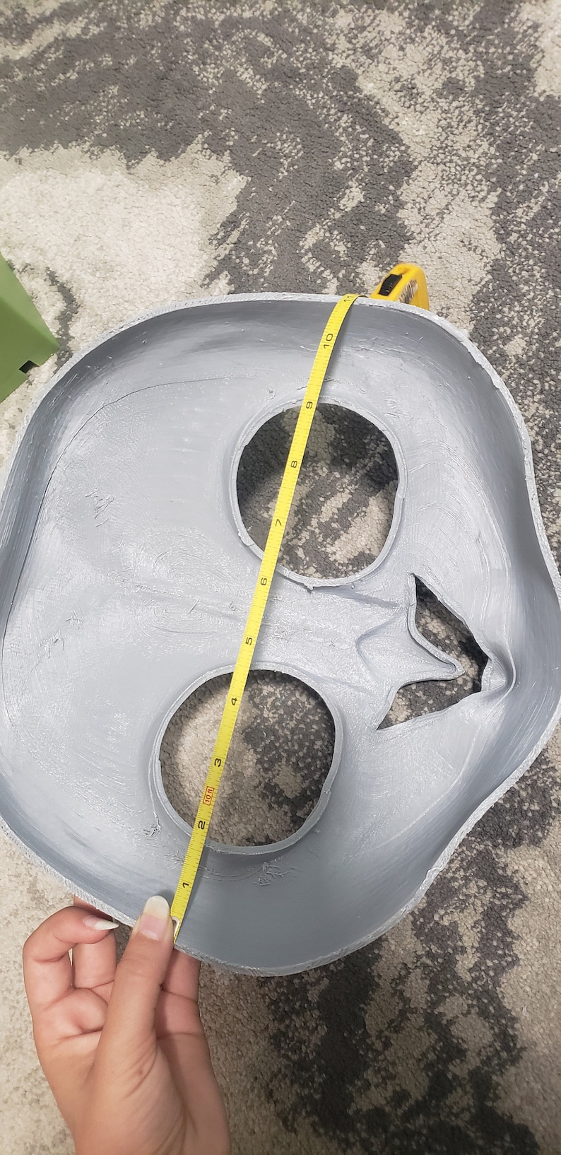 May include: A gray plaster mold of a face with two eye holes and a mouth hole. A yellow measuring tape is wrapped around the mold, measuring 10 inches.