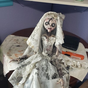 K&ouml;nnte beinhalten: Eine handgefertigte, gruselige Puppe in einem Brautkleid. Die Puppe hat ein besch&auml;digtes Gesicht und tr&auml;gt ein zerrissenes wei&szlig;es Hochzeitskleid und einen Schleier. Die Puppe sitzt auf einem Tisch und hat ein verst&ouml;rtes Aussehen.