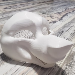 May include: A white, unpainted, furry animal mask with a large snout and open mouth. The mask is made of a smooth, hard material and has two eye holes.
