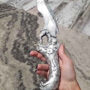 K&ouml;nnte beinhalten: Ein silberfarbener Dolch mit geschwungener Klinge und kunstvollem Griff. Der Griff weist ein detailliertes, skulpturales Design auf. Der Dolch wird in einer Hand gehalten, was seine Gr&ouml;&szlig;e und Handwerkskunst zeigt. Die Klinge scheint scharf zu sein.