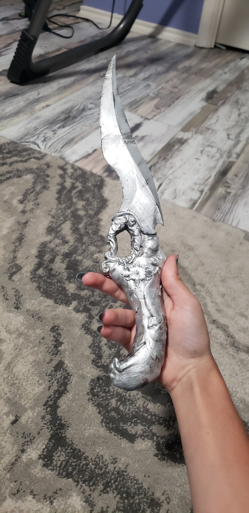 K&ouml;nnte beinhalten: Ein silberfarbener Dolch mit geschwungener Klinge und kunstvollem Griff. Der Griff weist ein detailliertes, skulpturales Design auf. Der Dolch wird in einer Hand gehalten, was seine Gr&ouml;&szlig;e und Handwerkskunst zeigt. Die Klinge scheint scharf zu sein.