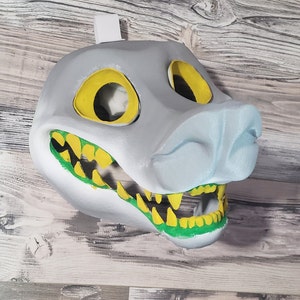 K&ouml;nnte beinhalten: Eine grau-stilisierte Maske mit gelben Augen&ouml;ffnungen und einer hellblauen Schnauze. Der Mund ist ge&ouml;ffnet und zeigt gelbe Z&auml;hne und gr&uuml;nes Zahnfleisch. Die Maske ist zum Tragen bestimmt und hat oben eine wei&szlig;e Lasche zum Aufh&auml;ngen.
