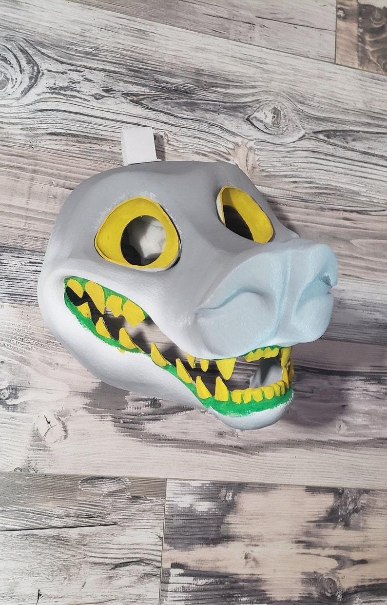 K&ouml;nnte beinhalten: Eine grau-stilisierte Maske mit gelben Augen&ouml;ffnungen und einer hellblauen Schnauze. Der Mund ist ge&ouml;ffnet und zeigt gelbe Z&auml;hne und gr&uuml;nes Zahnfleisch. Die Maske ist zum Tragen bestimmt und hat oben eine wei&szlig;e Lasche zum Aufh&auml;ngen.