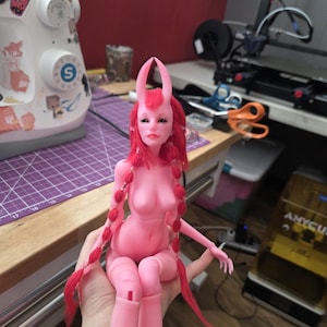 K&ouml;nnte beinhalten: Eine rosa Kunstpuppe mit roten Z&ouml;pfen und rosa H&ouml;rnern. Die Puppe sitzt mit angewinkelten Beinen. Sie hat einen ruhigen Gesichtsausdruck. Die Puppe ist ein einzigartiges handgefertigtes Produkt.