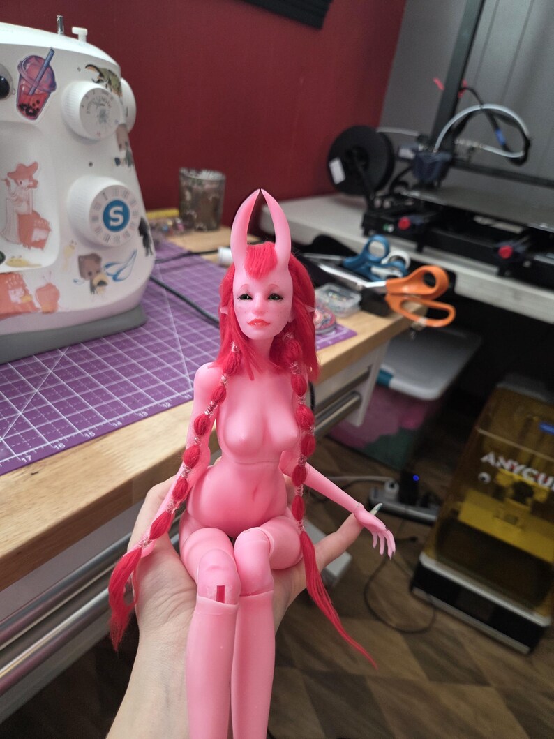 K&ouml;nnte beinhalten: Eine rosa Kunstpuppe mit roten Z&ouml;pfen und rosa H&ouml;rnern. Die Puppe sitzt mit angewinkelten Beinen. Sie hat einen ruhigen Gesichtsausdruck. Die Puppe ist ein einzigartiges handgefertigtes Produkt.